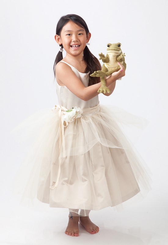 Mädchen im cremefarbenen Tüllkleid hält die Folkmanis Handpuppe Lustiger Frosch in olivgrünem Plüsch in die Kamera.