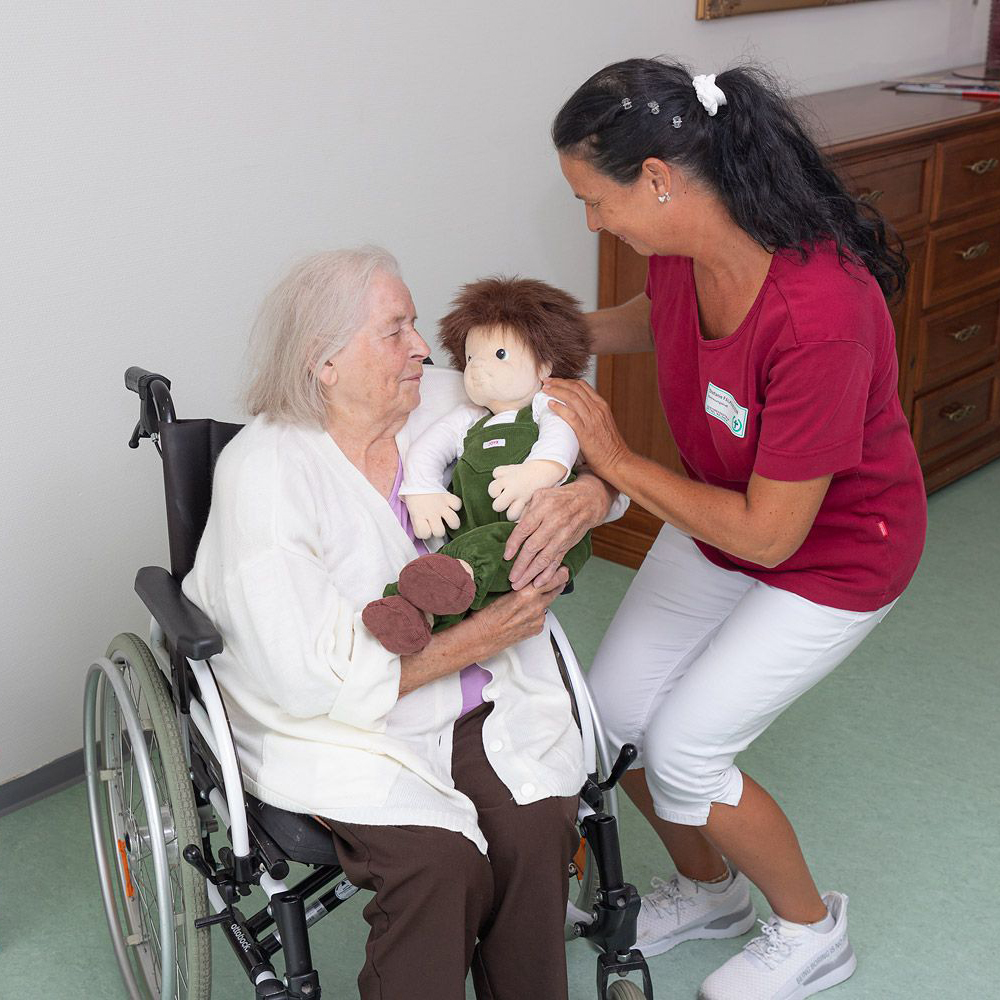 Caregiver handing the Joyk empathy doll Marco to a senior lady in a wheelchair