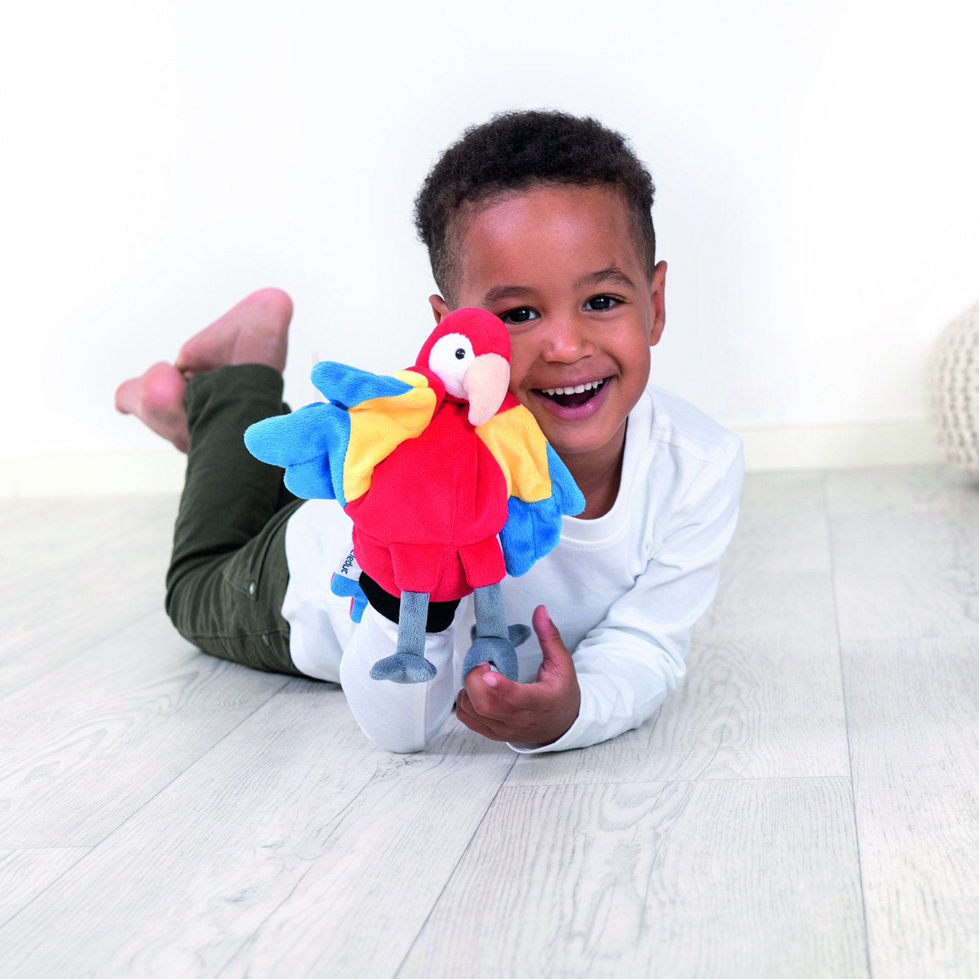 Boy smiling and lying on the floor with the Beleduc hand puppet parrot, usage example