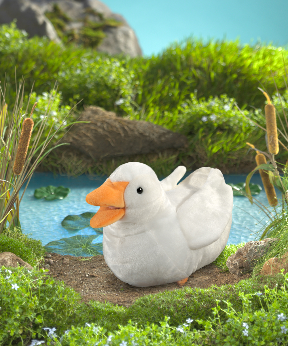 Folkmanis duck hand puppet in white with an orange beak, sitting in front of a pond with lily pads and reeds