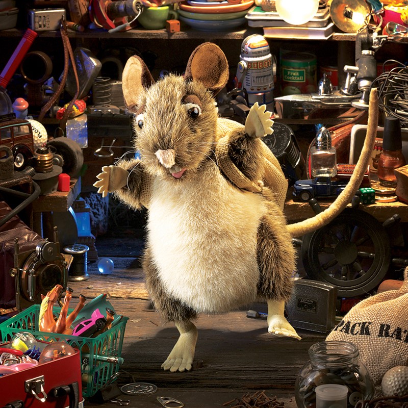 Folkmanis Handpuppe Packratte mit graubraunem Fell und Jutesack steht aufrecht in einer Kulisse voller Fundstücke