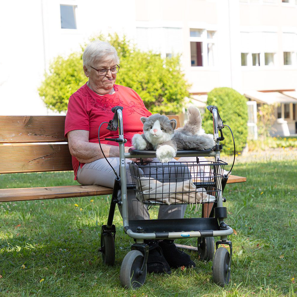 Die Joyk Empathiepuppe Billy liegt auf einem Rollator neben einer Seniorin im Garten