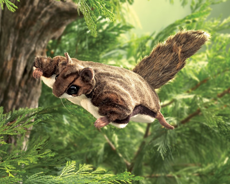 Folkmanis Handpuppe Flughörnchen in braunem Plüsch, in Gleitpose zwischen grünen Tannenzweigen fotografiert