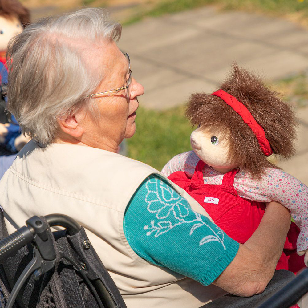 Seniorin hält die Joyk Empathiepuppe Ingrid im Arm im Garten