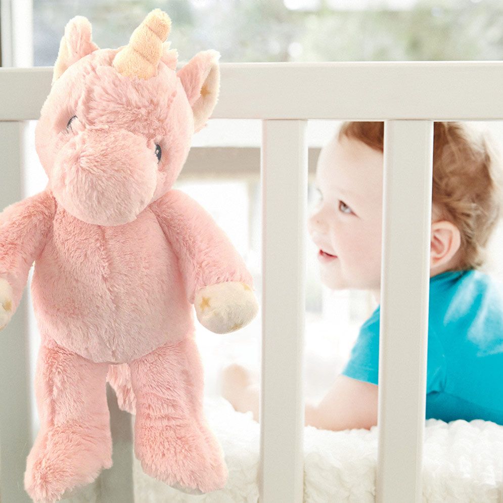 Cloud b Einschlafhilfe Ella Einhorn, Anwendungsfoto: rosa Kuscheltier hängt am Gitterbett, Baby im Hintergrund