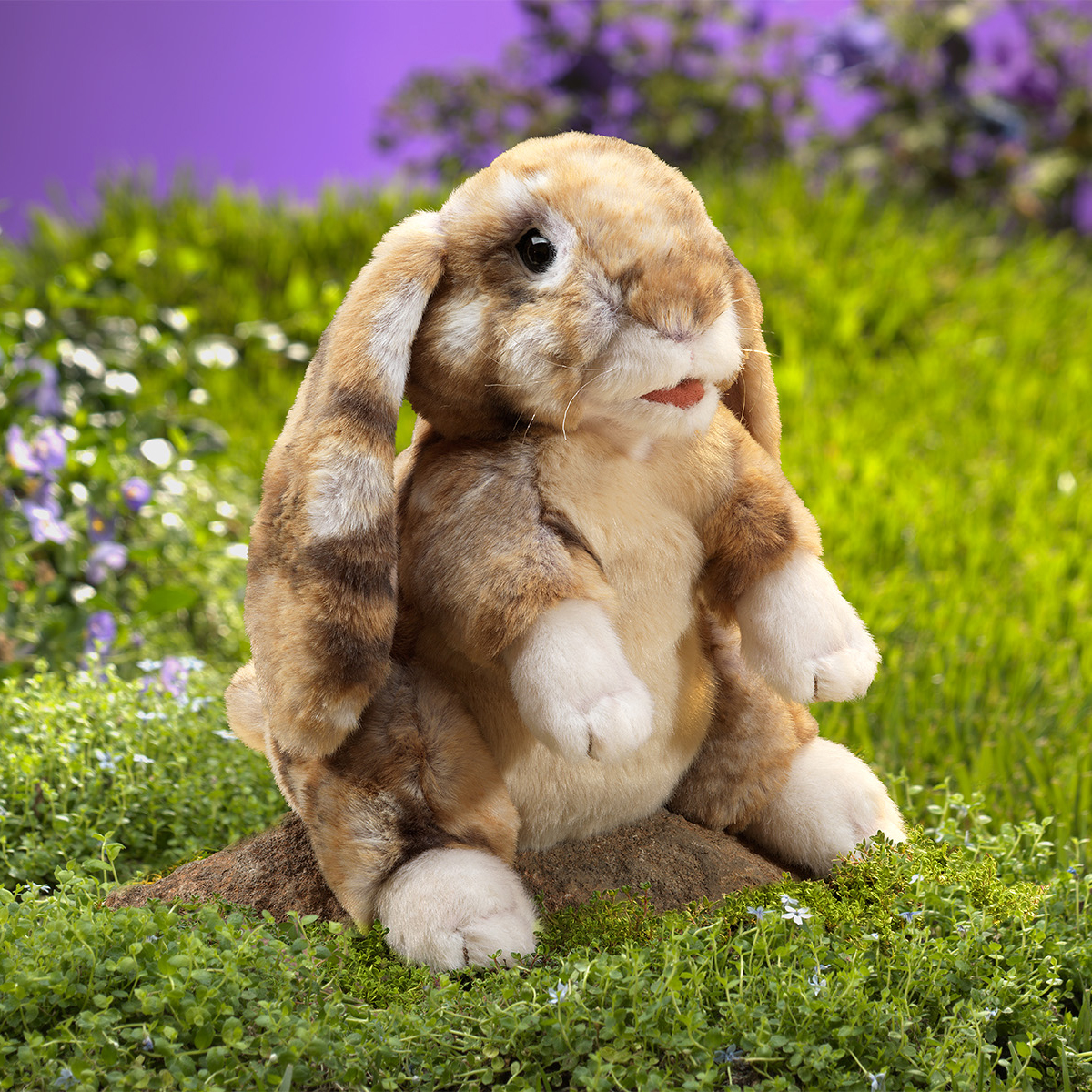 Folkmanis hand puppet lop-eared rabbit in brown tones, sitting upright with drooping ears on a clump of earth in front of a green meadow