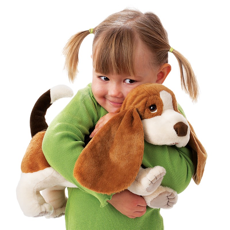 Kleines Mädchen hält die Folkmanis Handpuppe Basset mit langen Schlappohren und braun-weißem Fell im Arm.