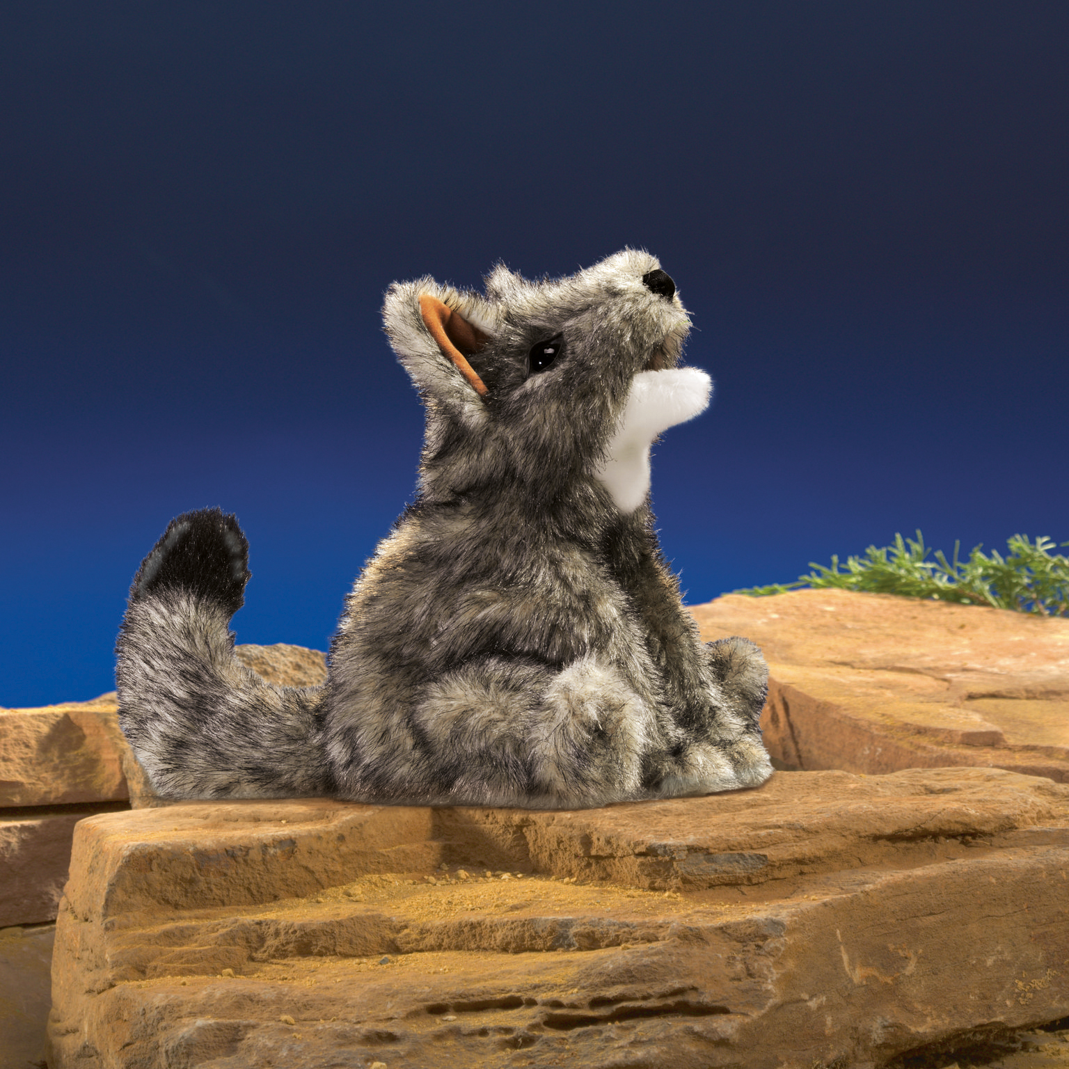 Folkmanis hand puppet small coyote with grey-melange fur, white throat and raised head, sitting on sandstone rocks