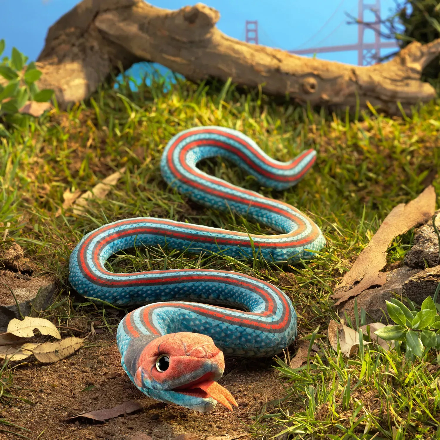 Folkmanis Fingerpuppe Strumpfbandnatter in Blau-Rot mit geöffnetem Maul, in gewundener Pose auf Waldboden mit Ast im Hintergrund