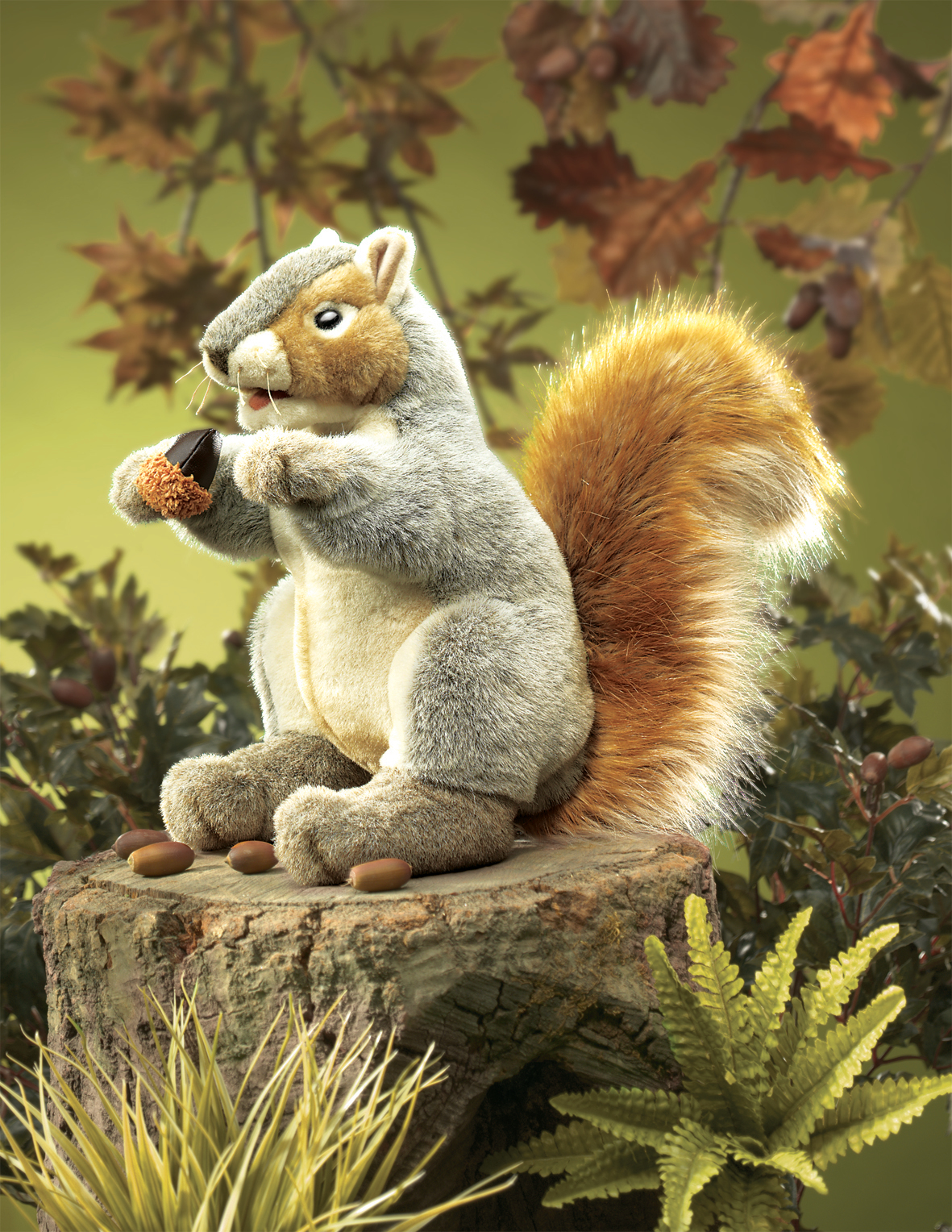 Folkmanis grey squirrel hand puppet with an orange tail holding an acorn, sitting on a tree stump against an autumn backdrop