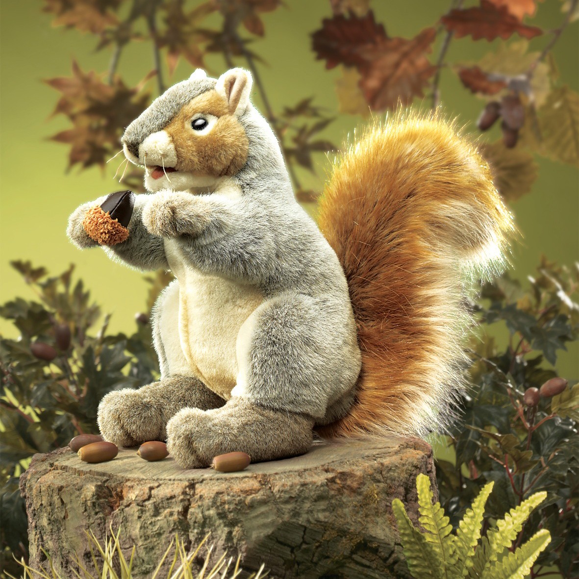 Folkmanis grey squirrel hand puppet with an orange tail holding an acorn, sitting on a tree stump against an autumn backdrop