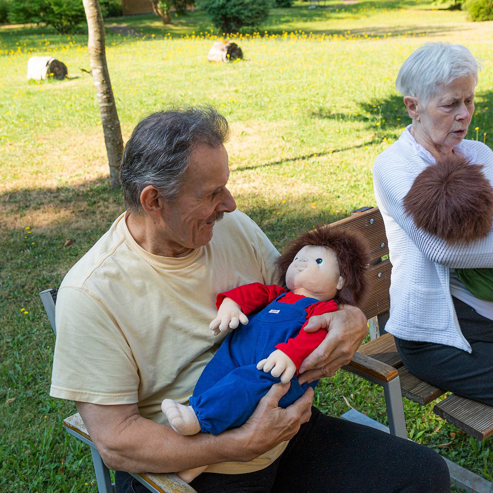 Senior hält die Joyk Empathiepuppe Johan im Arm bei einer Gartenrunde