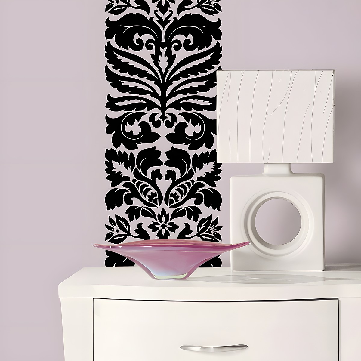 RoomMates wall decal Class A Peel & Stick, black damask ornament on a light lilac wall behind a chest of drawers with a lamp and bowl