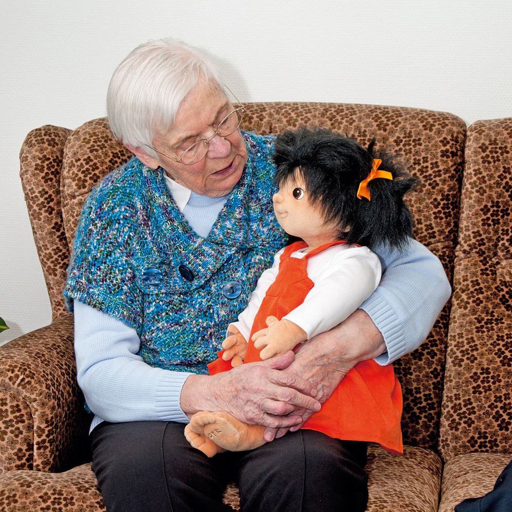 Seniorin mit der Joyk Empathiepuppe Emelie auf dem Schoß im Sessel