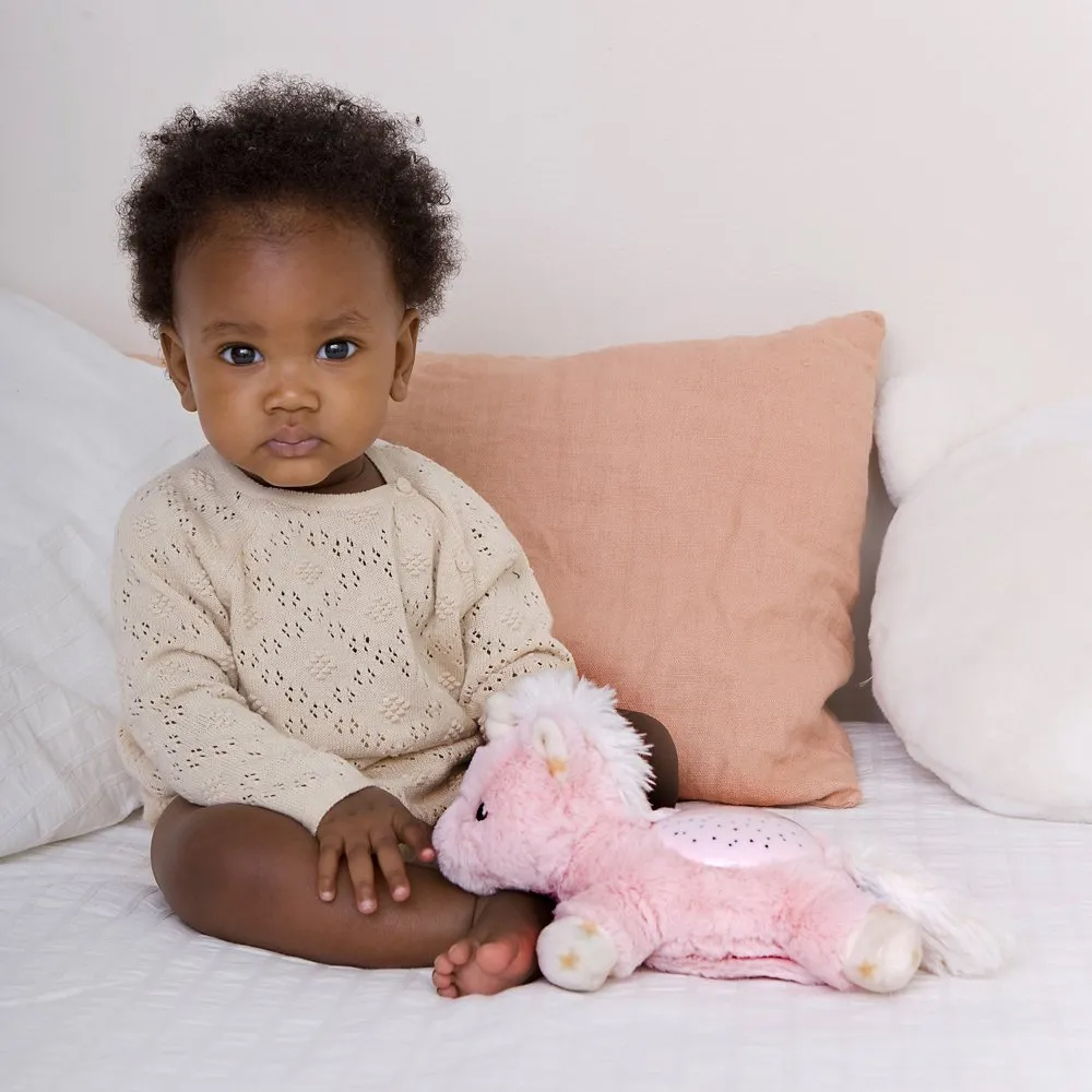 Cloud b Einschlafhilfe Dream Buddies Einhorn, Anwendungsfoto: Baby sitzt im Bett neben rosa Kuscheltier