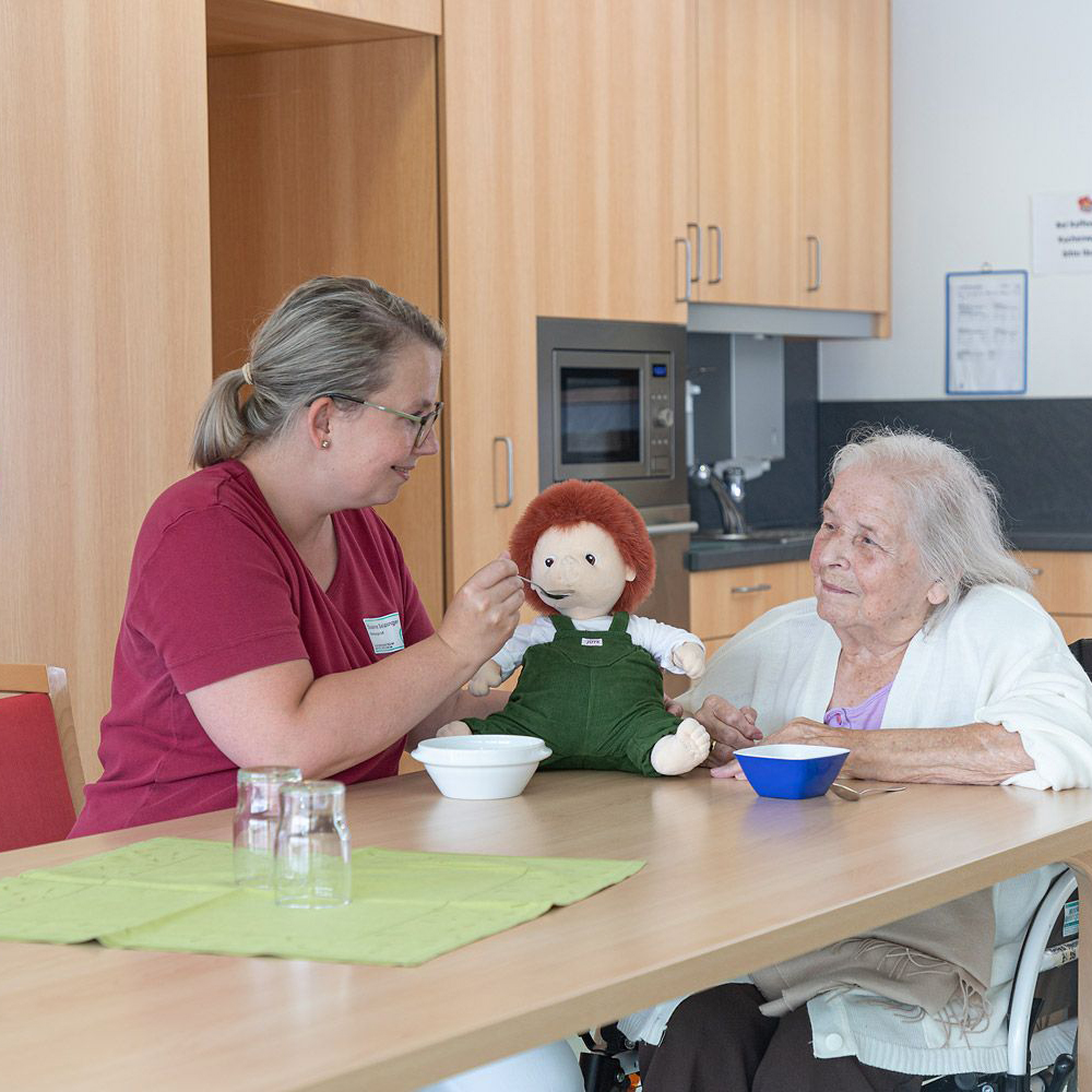 Caregiver and senior lady with the Joyk empathy doll Sam sharing a meal at the table