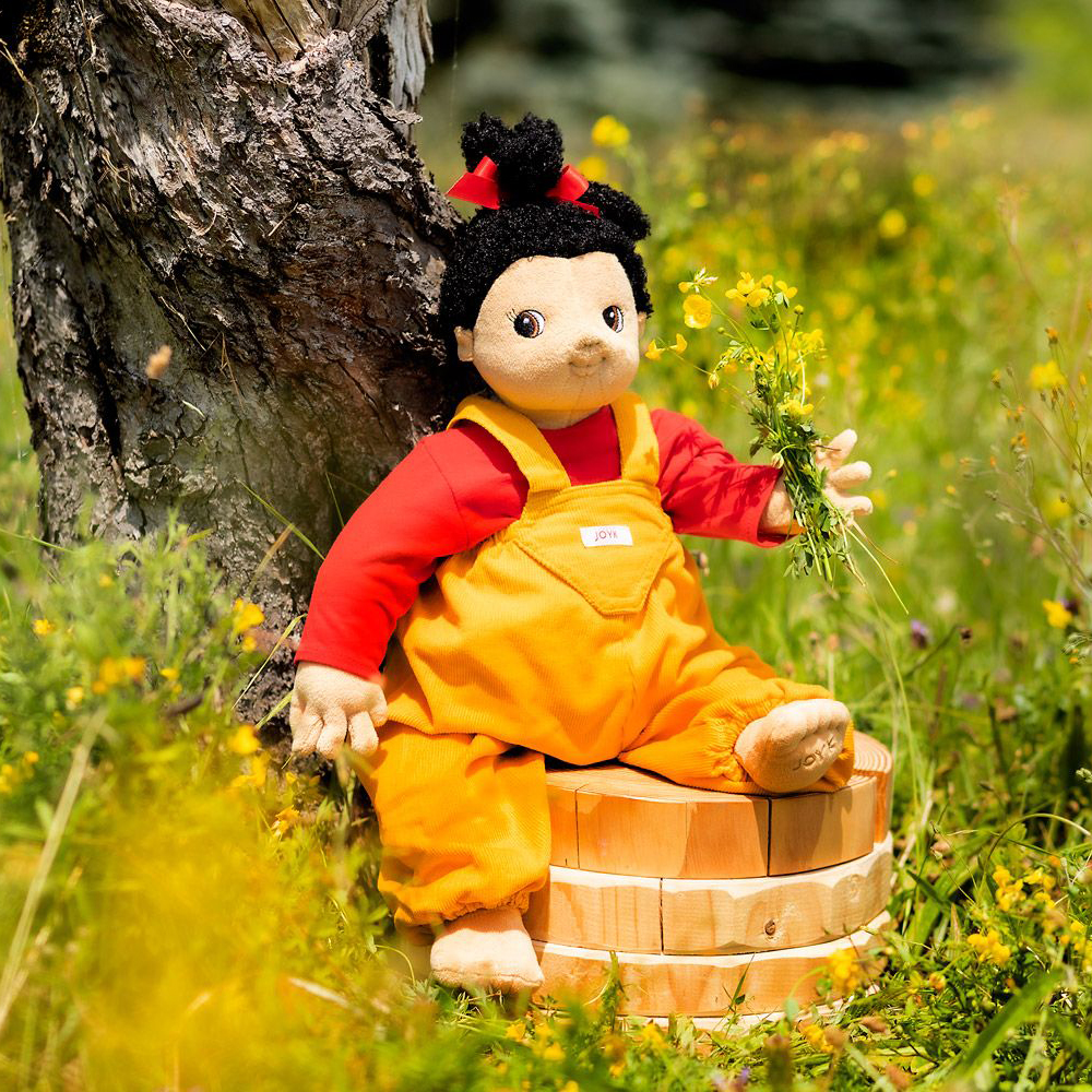 Joyk Empathiepuppe Naomi sitzt auf einem Holzbottich in einer Blumenwiese