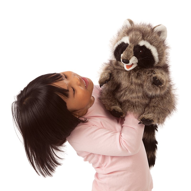 Mädchen hält Folkmanis Handpuppe Waschbär mit geöffnetem Maul in die Höhe, grau-braunes Fell mit schwarzer Gesichtsmaske.