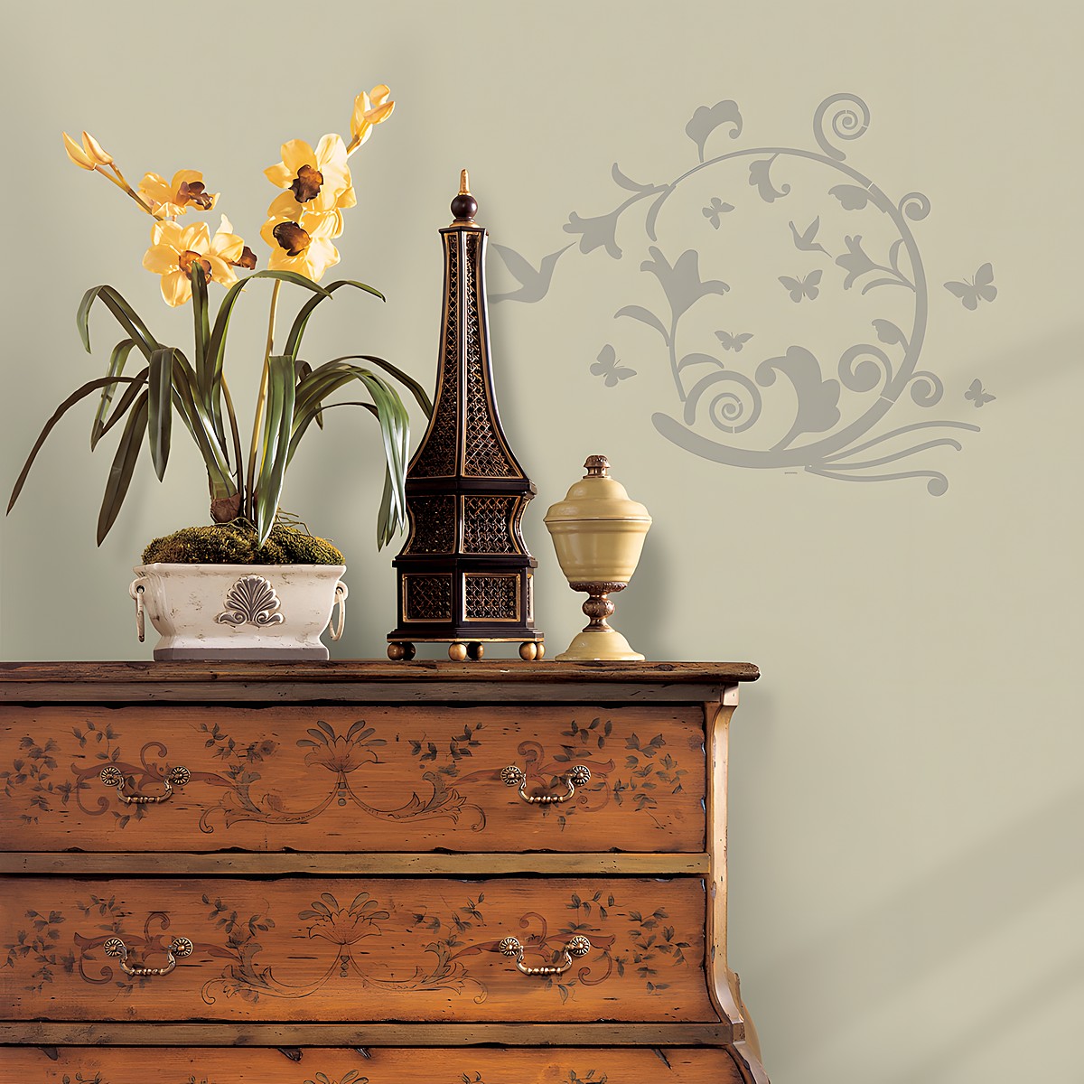 RoomMates wall decal Graphic Oval Peel & Stick, silver-coloured oval vine ornament with butterflies on a grey-green wall above a chest of drawers