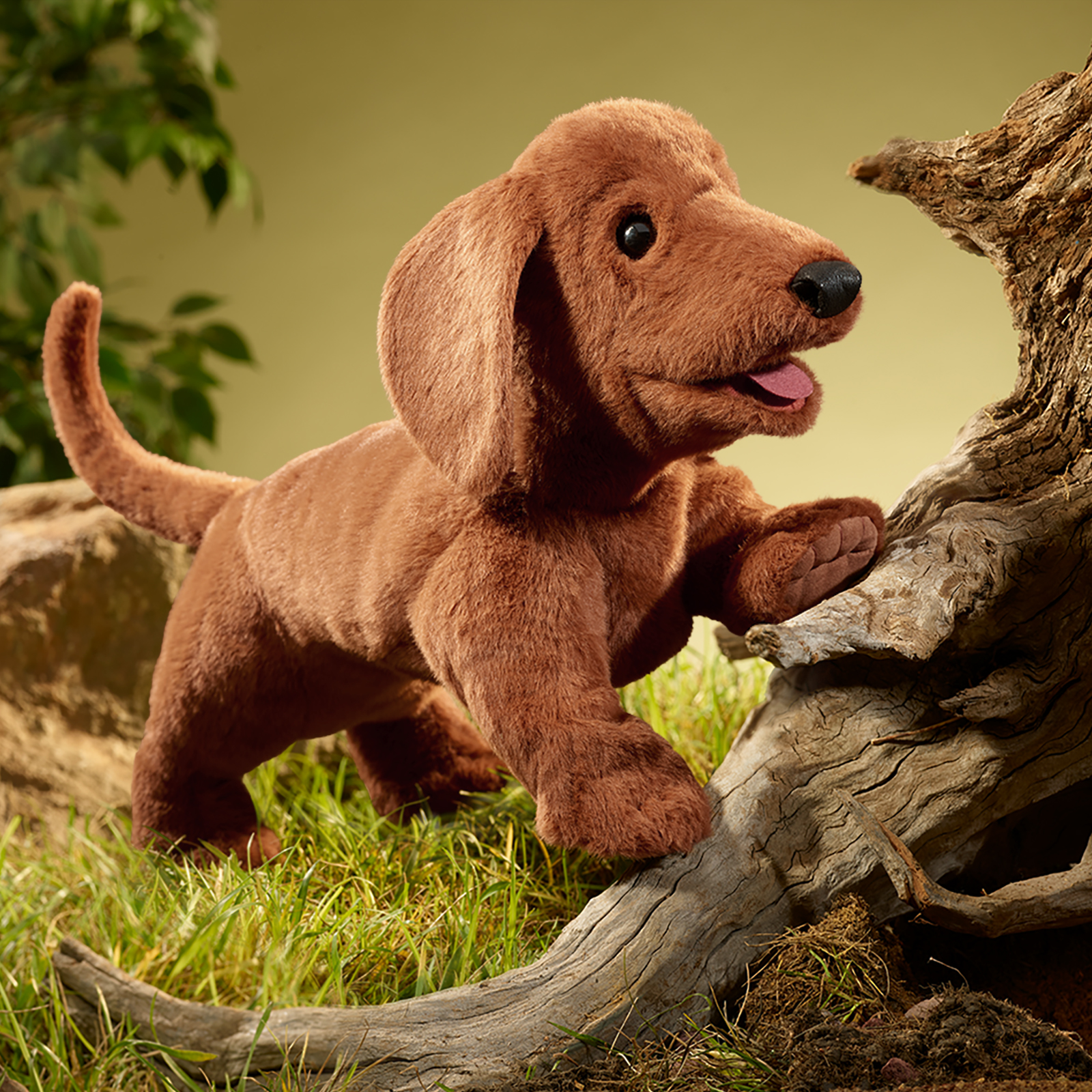 Folkmanis Handpuppe Dackel stützt eine Vorderpfote auf einen Ast, das Maul geöffnet, im Hintergrund Gras und Laub.