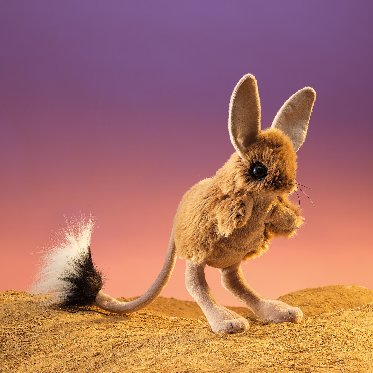 Folkmanis jerboa hand puppet with long ears, black-and-white tail and sand-coloured fur, posed upright on a sand dune