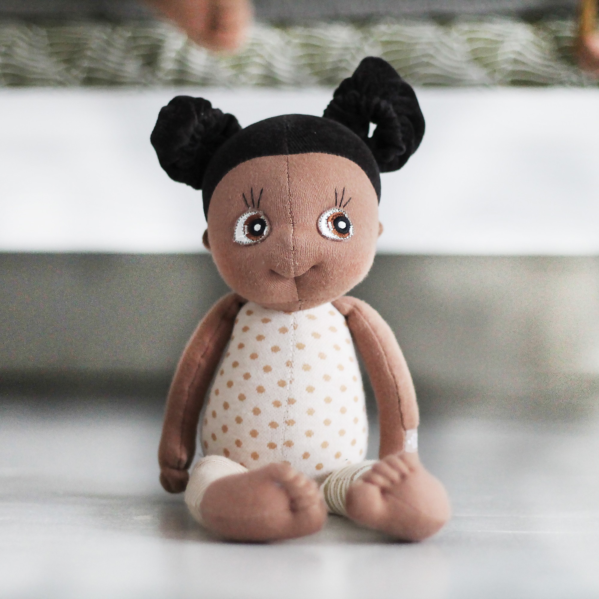 Rubens EcoBuds Mini-Puppe Flora sitzt mit gepunktetem Top vor einem Bett