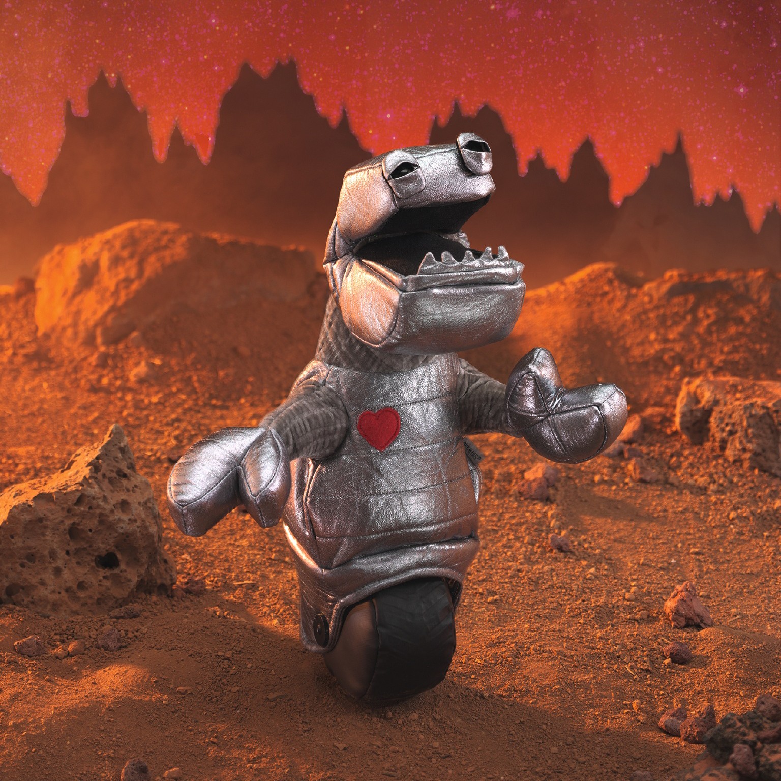 Folkmanis robot hand puppet in a silver full-body suit with a red heart on the chest, open mouth and outstretched claw hand, against a Mars-like backdrop