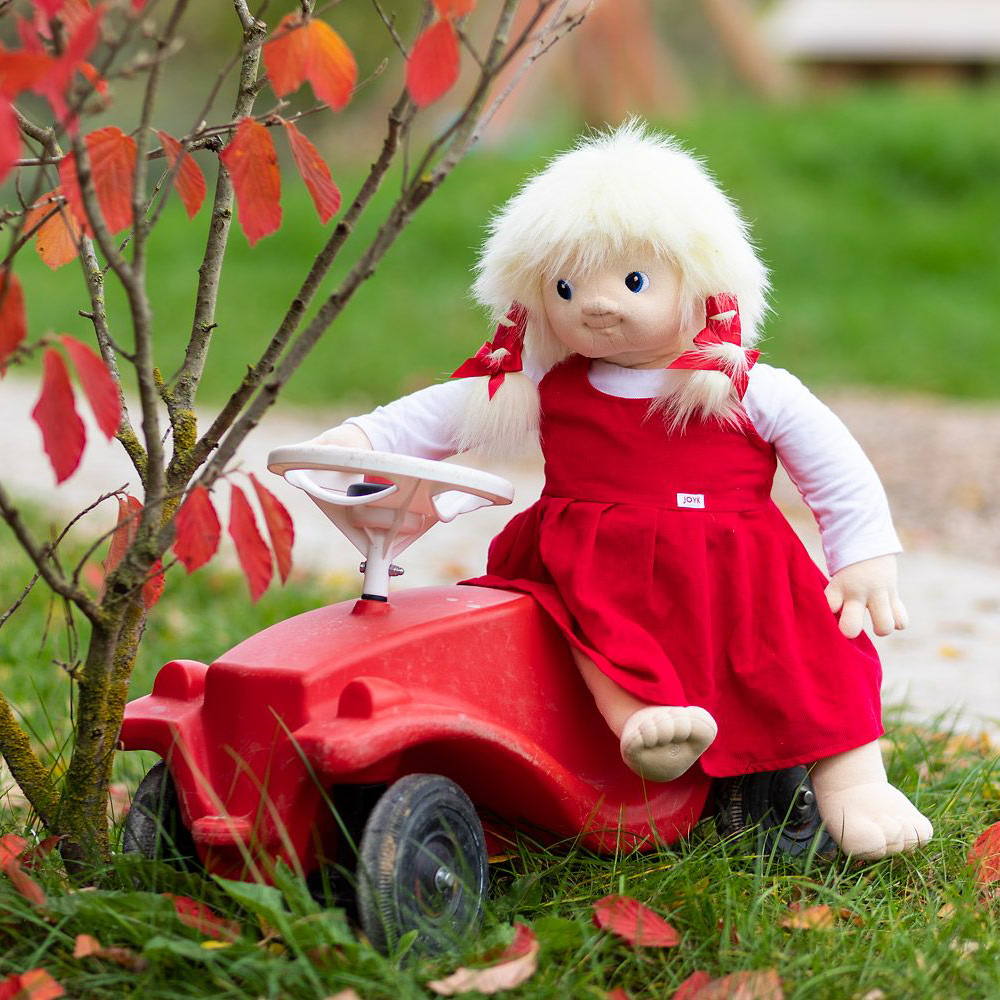 Die Joyk Empathiepuppe Nelly sitzt auf einem roten Rutschauto im Herbstgarten