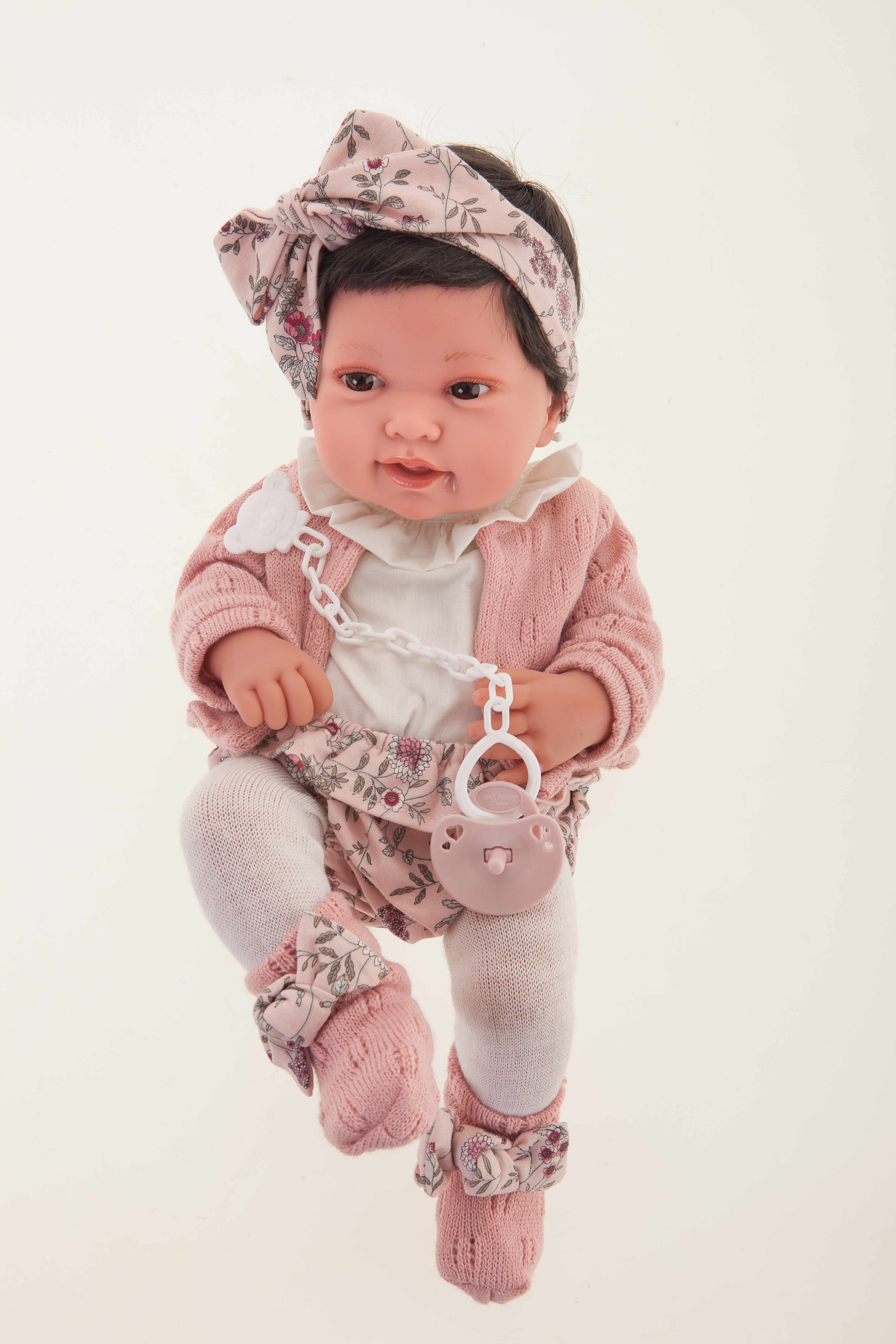 Antonio Juan Babypuppe Pipa, realistische Puppe mit dunklem Haar, Blumen-Stirnband und rosa Strickjacke, Frontalansicht mit weißen Strumpfhosen