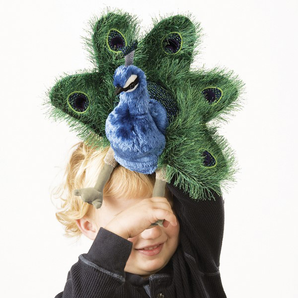 Kind hält Folkmanis Handpuppe kleiner Pfau vor sein Gesicht, mit blauem Gefieder und grünem Rad aus Pfauenaugen-Federn.