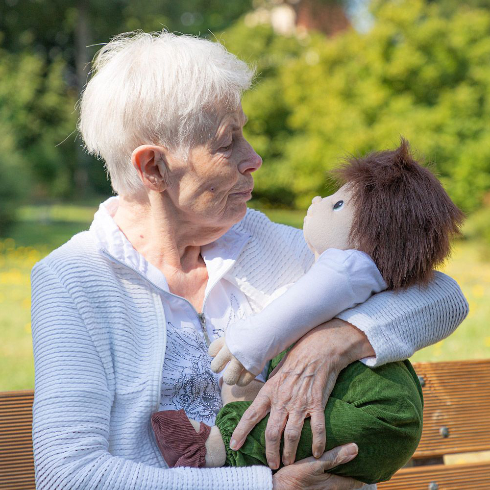 Senior lady holding the Joyk empathy doll Marco in her arms on a park bench