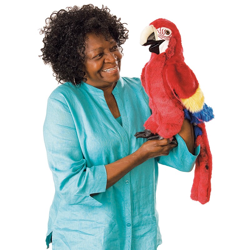 Folkmanis Handpuppe Ara in leuchtendem Rot mit gelb-blauen Flügeln, von einer lächelnden Frau auf dem Arm getragen
