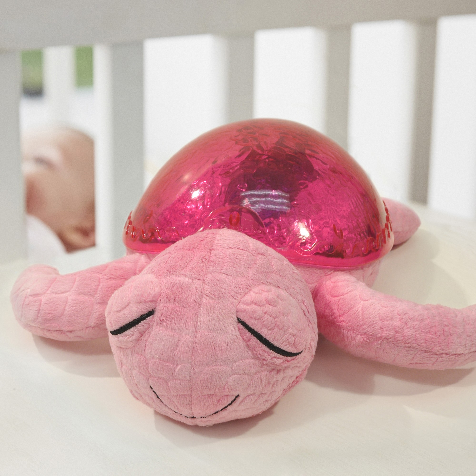 Cloud b Einschlafhilfe Tranquil Turtle Pink, Kuscheltier am Gitterbett mit schlafendem Baby im Hintergrund