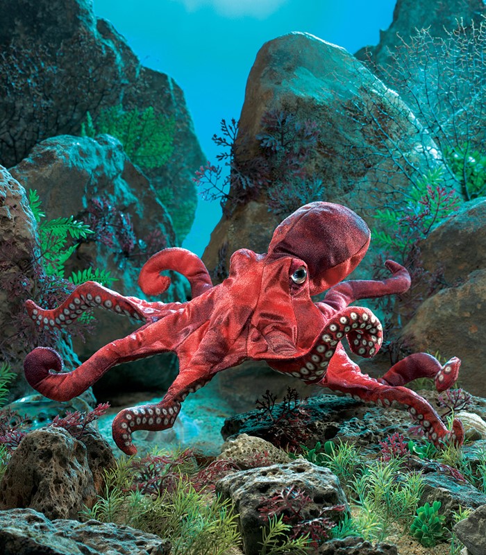 Folkmanis hand puppet red octopus with white suckers on eight tentacles, set against a staged ocean floor with rocks and seaweed