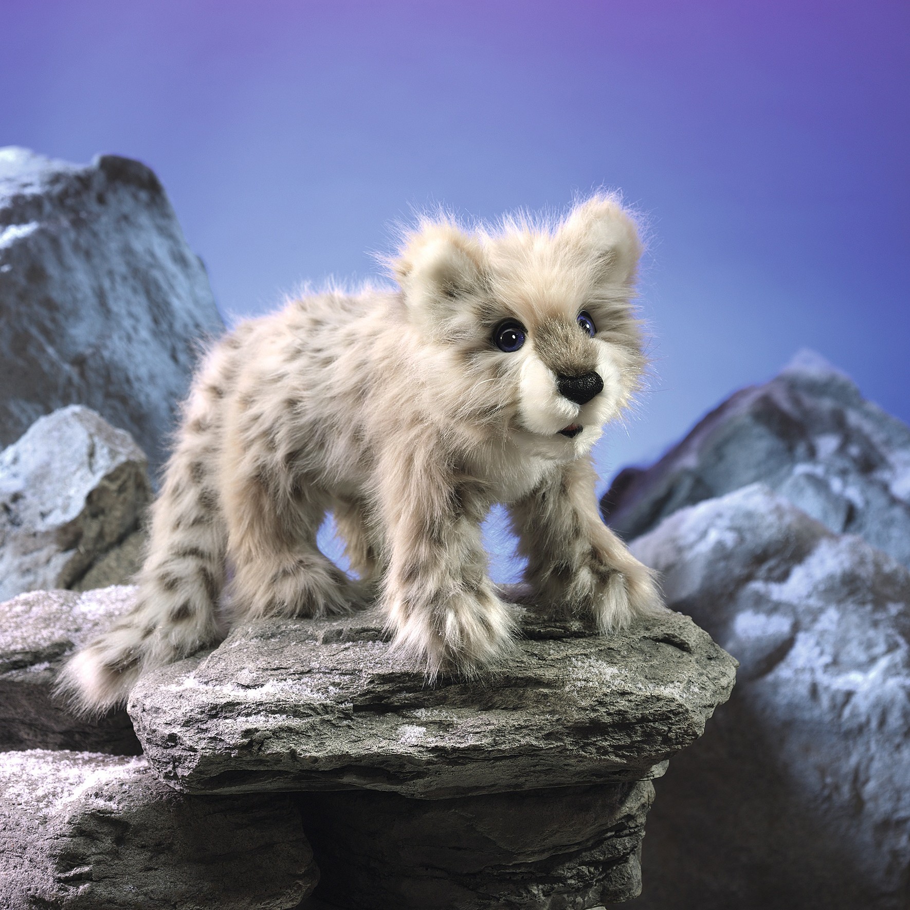 Folkmanis Handpuppe Baby Schneeleopard mit grau-beigem Fleckenfell, stehend auf einem Felsen vor blau-violettem Hintergrund
