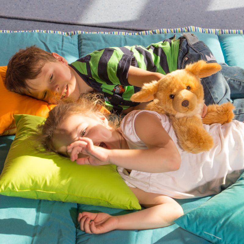 Zwei Kinder liegen auf Kissen und kuscheln mit dem Joyk-Hund Brando